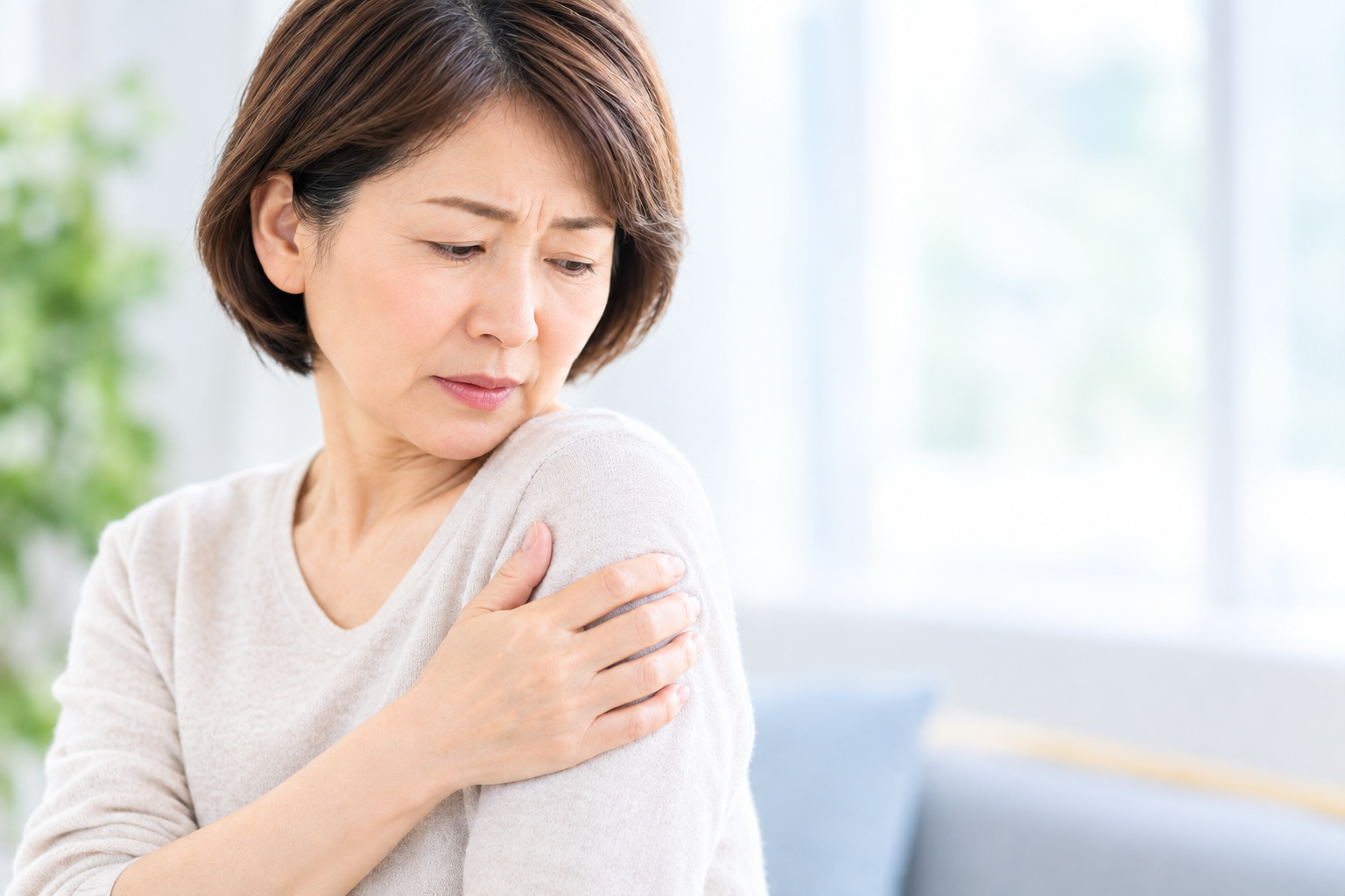 Middle-aged woman touching her shoulder in discomfort, looking down with a worried expression.